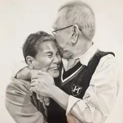 A highly detailed charcoal drawing on paper depicts an elderly couple in a loving embrace. The man, wearing glasses and a vest, gently kisses the woman’s forehead while holding her close. The woman smiles joyfully, her eyes crinkling with happiness. The artwork captures deep emotion and fine details in black and white.