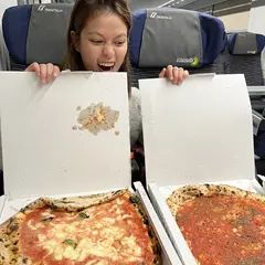 A young woman with light skin and brown hair excitedly holds open two pizza boxes containing Neapolitan-style pizzas. One pizza has melted mozzarella and tomato sauce, while the other is a simple marinara. She is sitting in a train with dark blue seats in the background, looking delighted at the food.